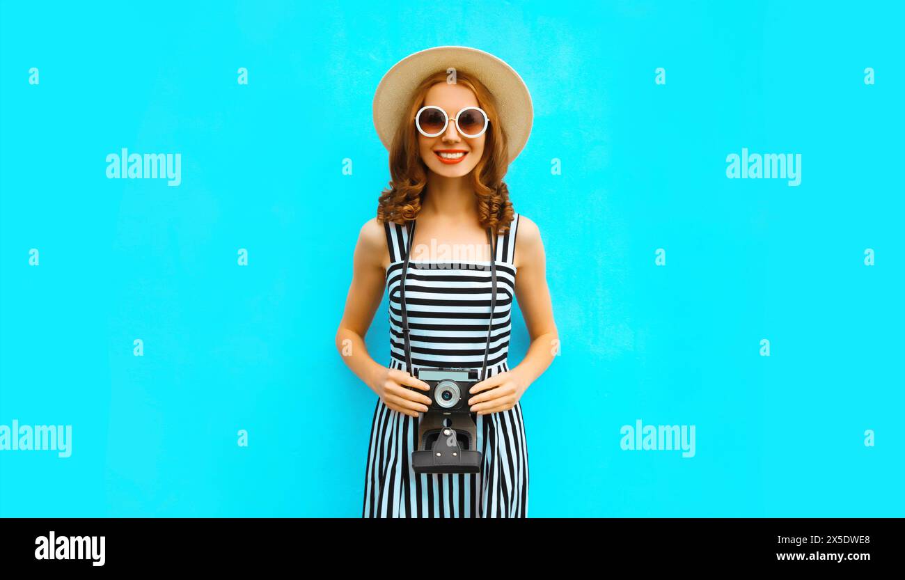 Portrait d'été de jeune femme heureuse souriante photographe avec caméra de film vintage portant chapeau de paille, robe, lunettes de soleil sur fond bleu Banque D'Images