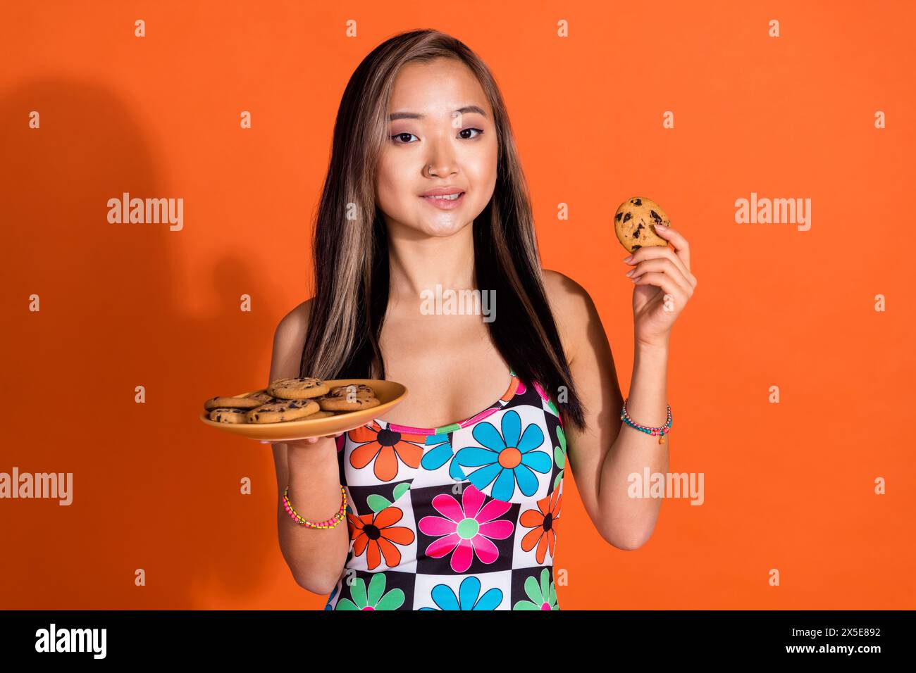 Photo de jolie femme adorable affamée porter une robe d'impression tenant un cookie savoureux dans les lèvres mortes de bras isolé sur fond de couleur orange Banque D'Images