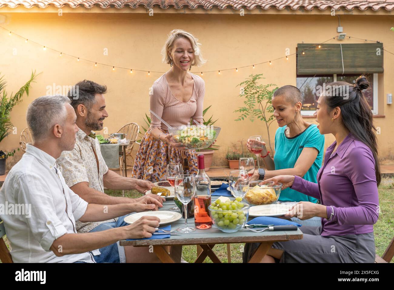 Amis multiethniques riant avec un grand sourire autour de la table. Femme sert la nourriture au patio de la maison. Sourire, gaieté concept. Banque D'Images