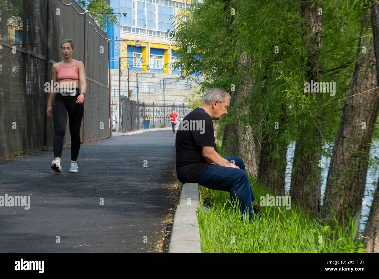 Un homme est assis sur un trottoir au bord de la rivière, une femme fait du jogging au bord de l'eau Banque D'Images