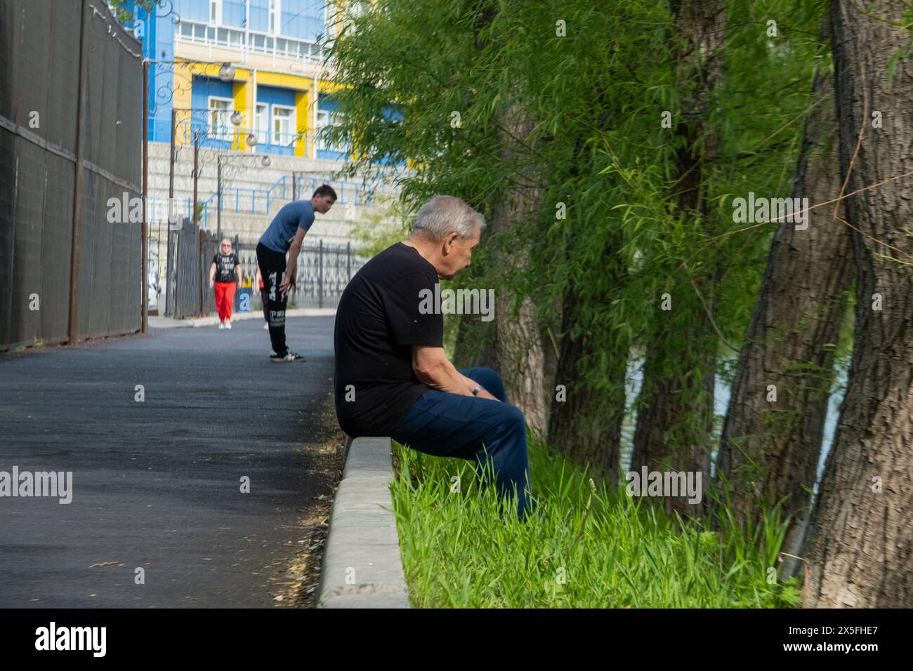 Un homme âgé est assis au bord de la route sur le trottoir et regarde tristement loin. Un jeune homme et une fille en arrière-plan Banque D'Images