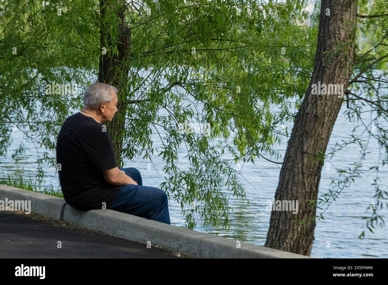 Un vieil homme en vêtements noirs est assis sur la rive au bord de l'eau et est triste de quelque chose. Vieillesse. Banque D'Images