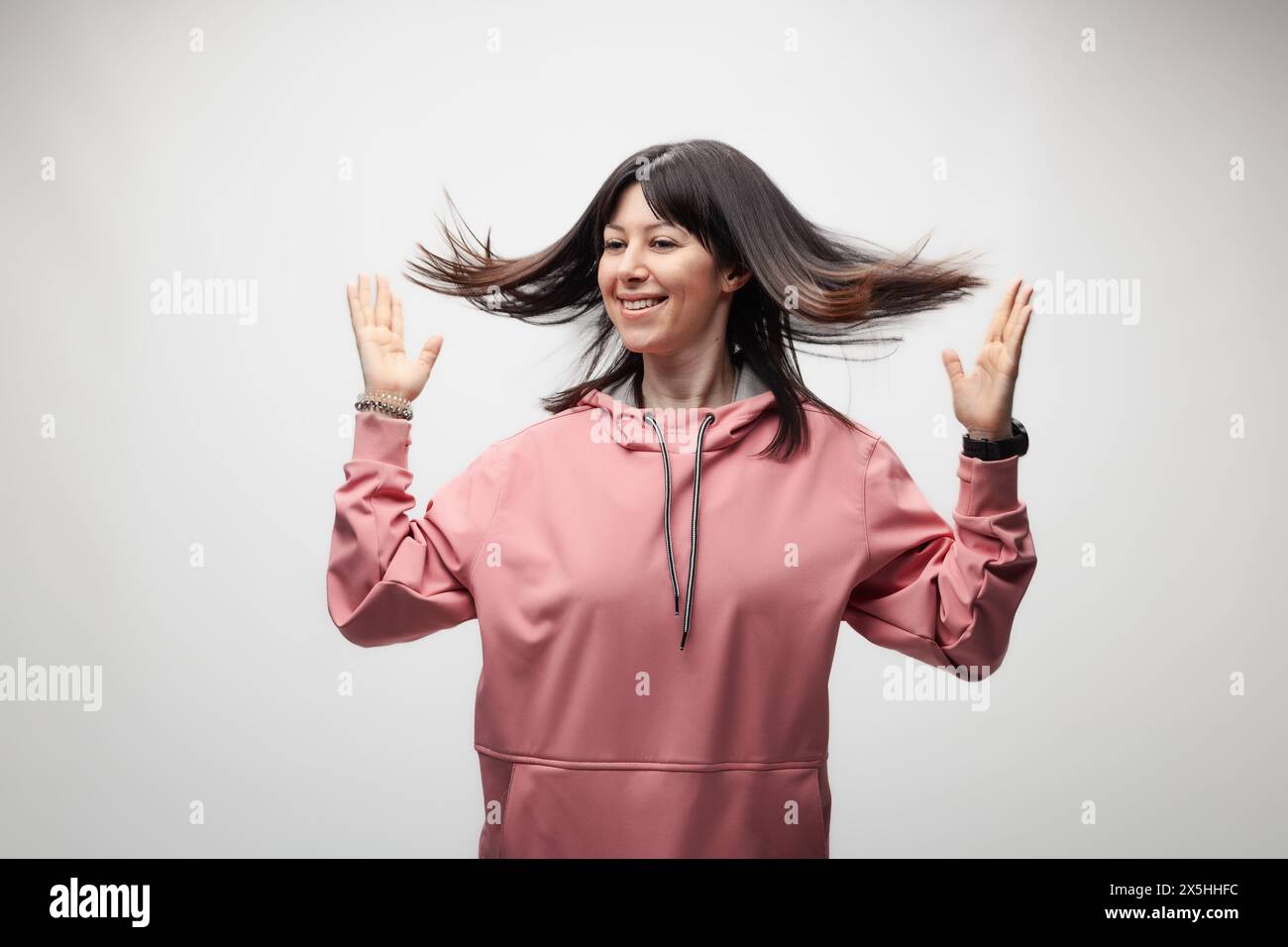 Une jeune femme joyeuse portant un sweat à capuche rose jette joyeusement ses cheveux, exprimant la liberté et le bonheur sur un fond clair. Banque D'Images