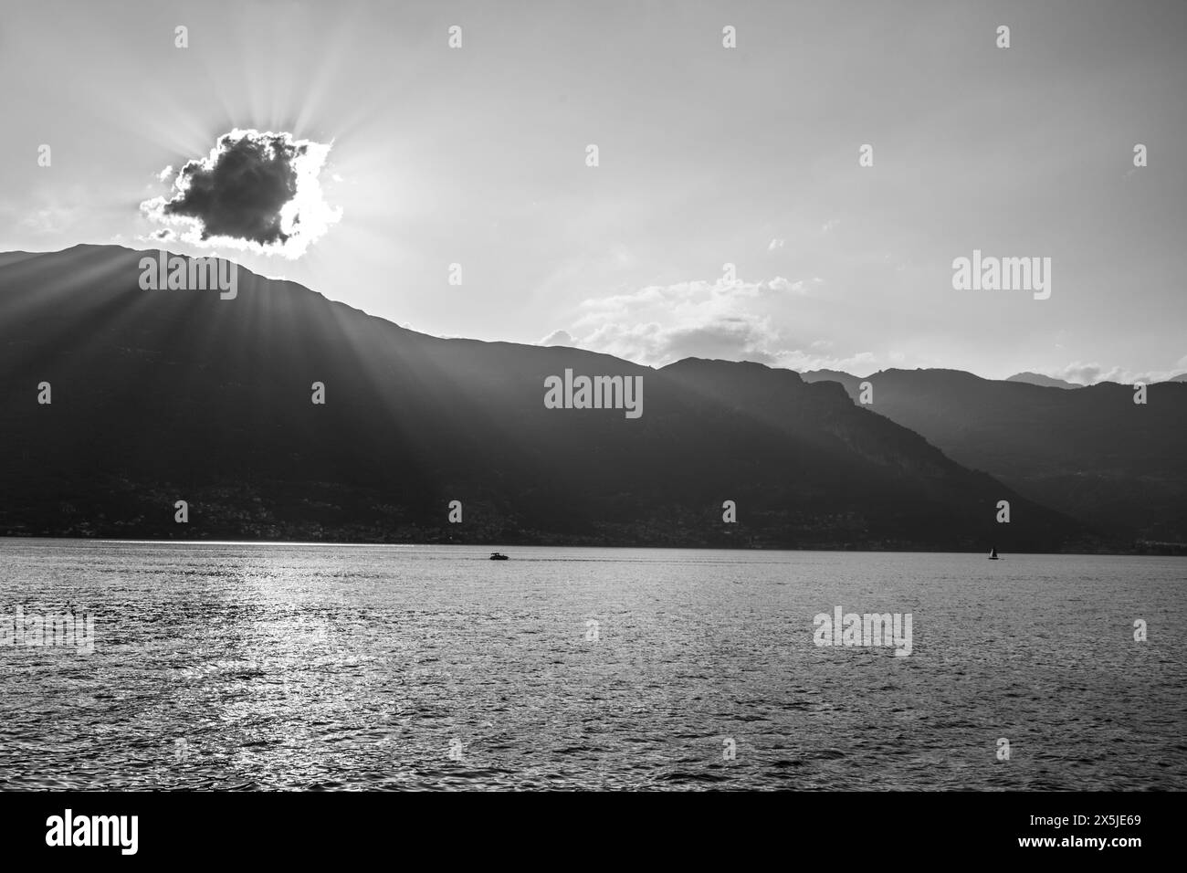 Une scène tranquille capturant la beauté sereine d'un lac de montagne au coucher du soleil. Les rayons du soleil traversent un nuage, projetant des faisceaux lumineux et des reflets étincelants sur la surface de l'eau. Un bateau solitaire profite de l'atmosphère paisible, entouré par la silhouette des montagnes. Image en noir et blanc. Banque D'Images