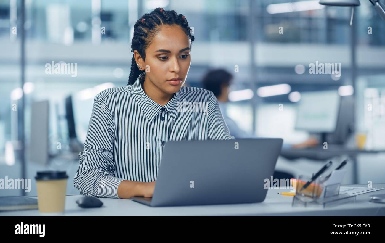 Bureau multi-ethnique : ingénieur INFORMATIQUE professionnel noir travaillant sur le bureau sur ordinateur portable. Femme d'affaires réussie et souriante avec des cheveux tressés fait un travail créatif. Concepteur de produit confiant Banque D'Images
