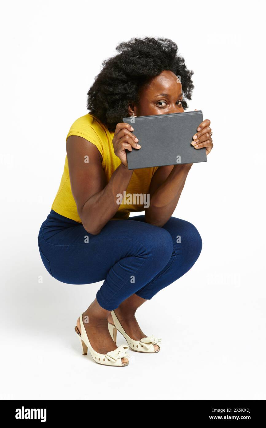 Photo de studio d'une femme accroupie et tenant un carnet devant le visage Banque D'Images