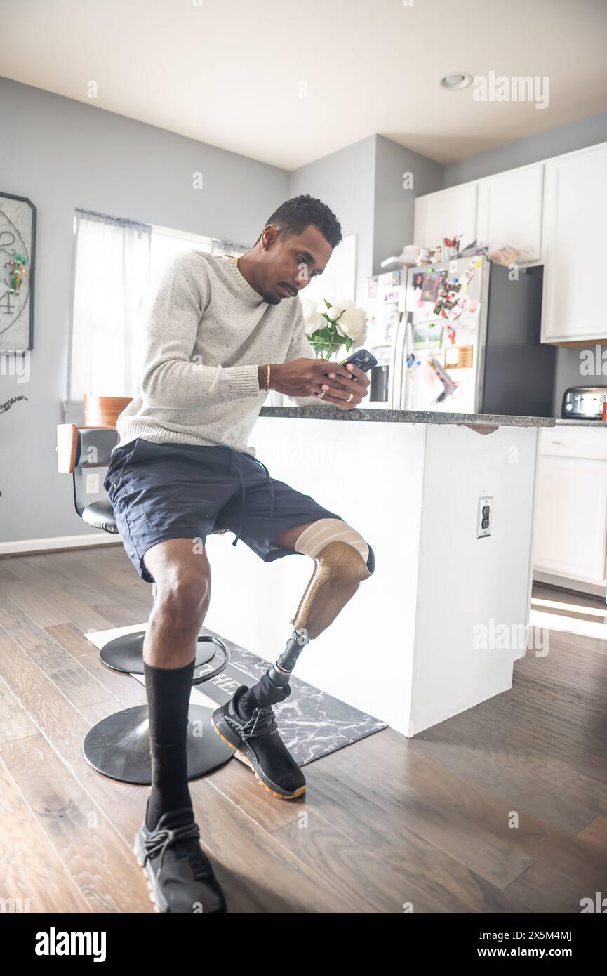 Homme avec une jambe prothétique utilisant un téléphone intelligent dans la cuisine Banque D'Images