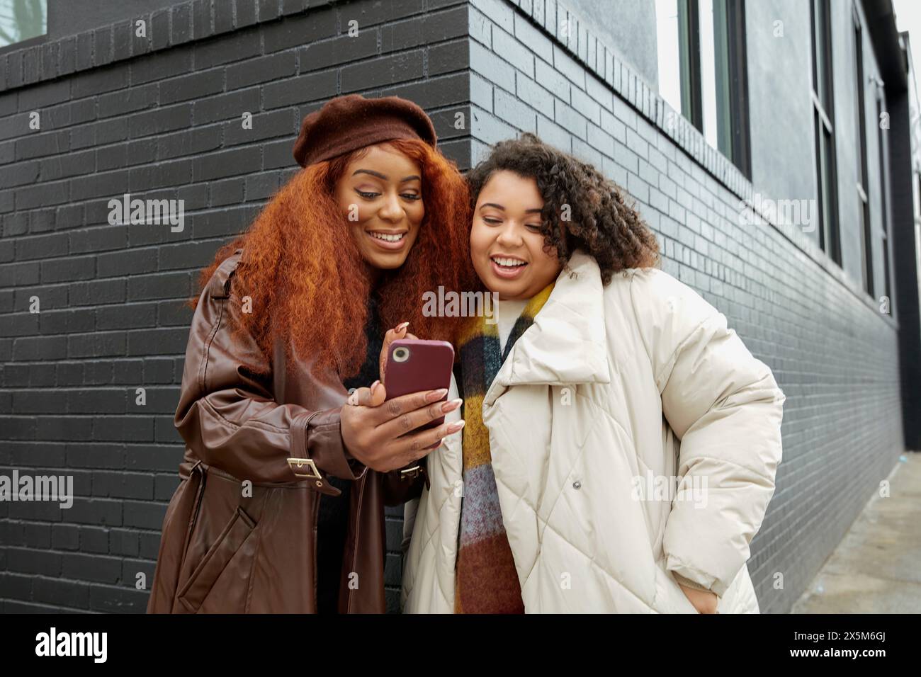 Des femmes souriantes regardant un téléphone intelligent devant un bâtiment noir Banque D'Images