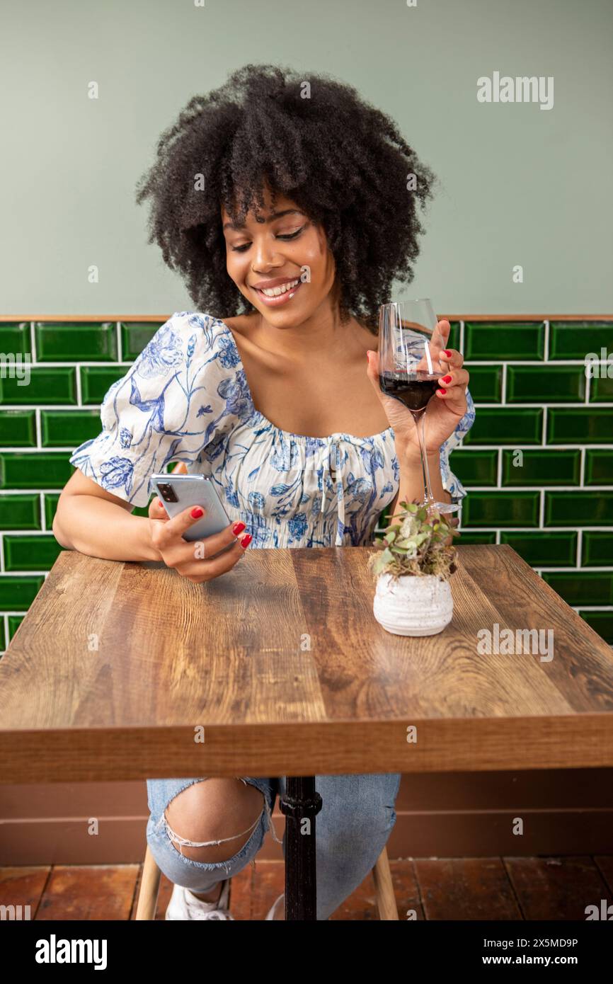 Royaume-Uni, Londres, femme souriante avec verre de vin rouge et téléphone intelligent à la table de café Banque D'Images