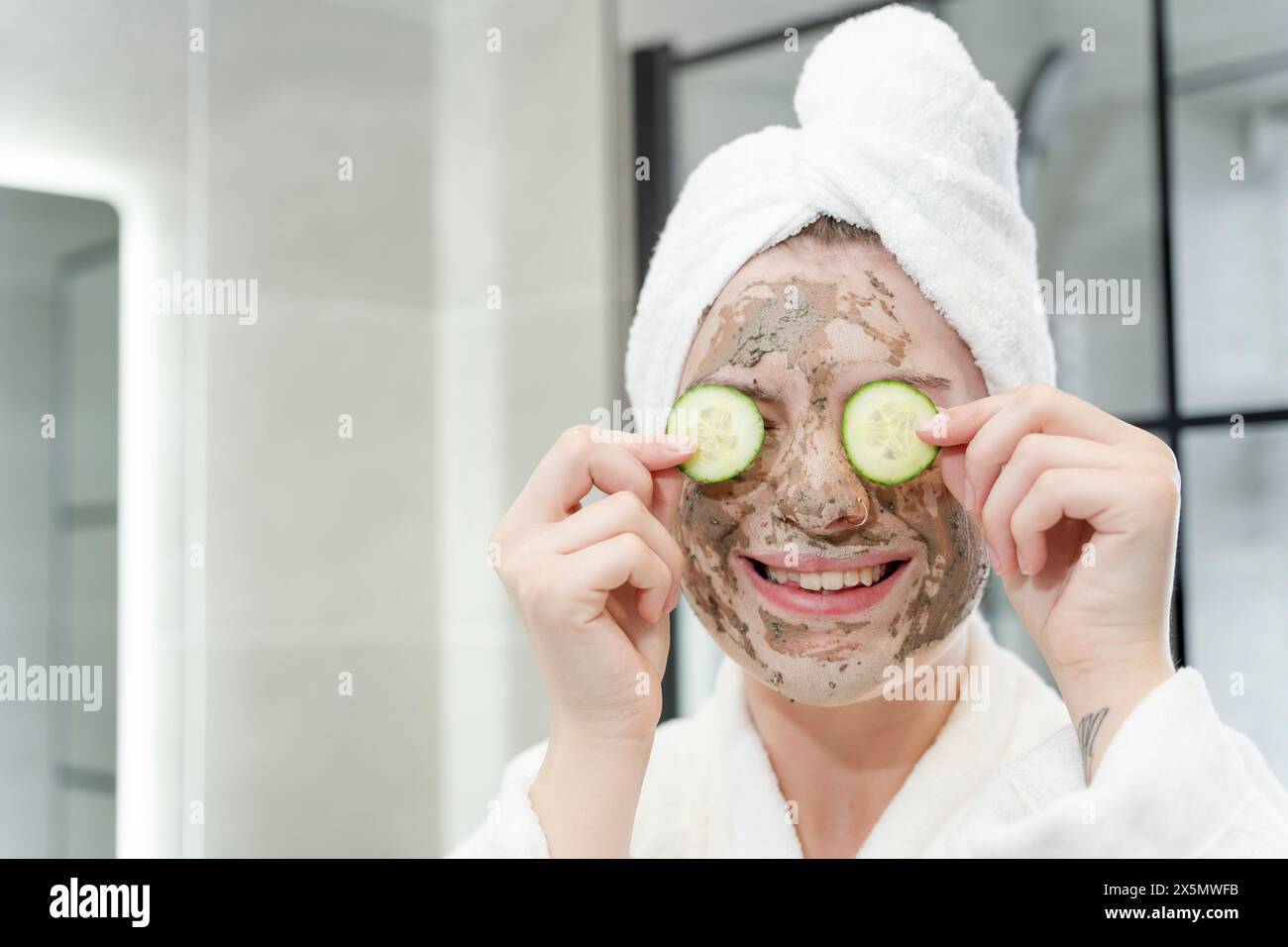 Femme souriante avec masque facial et tranches de concombre dans la salle de bain Banque D'Images