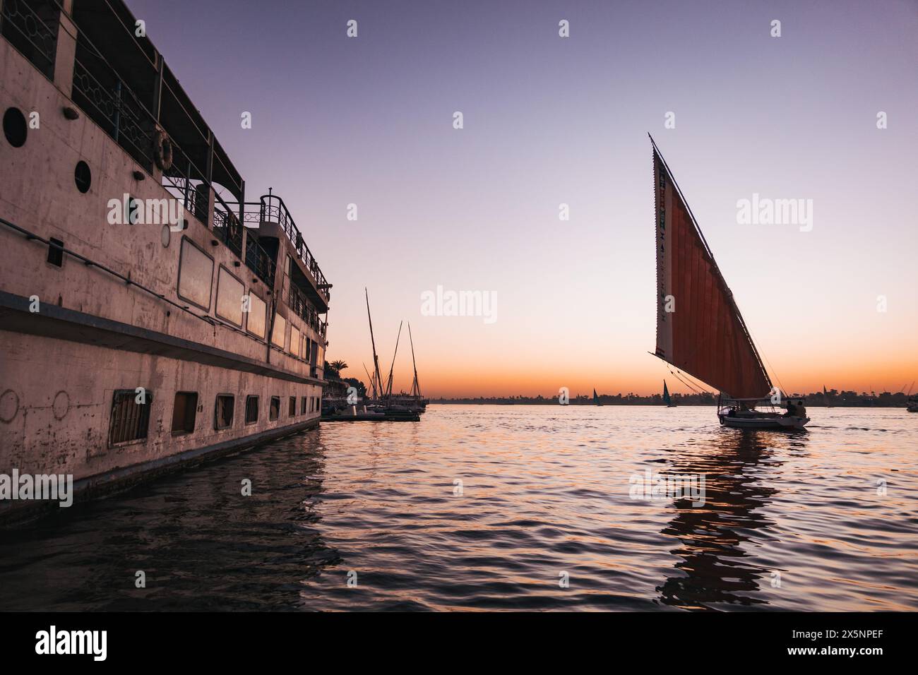 Une felouque navigue devant un bateau de croisière amarré sur le Nil au coucher du soleil, avec une eau calme et un ciel orange et violet doux au-dessus de l'horizon lointain. Banque D'Images
