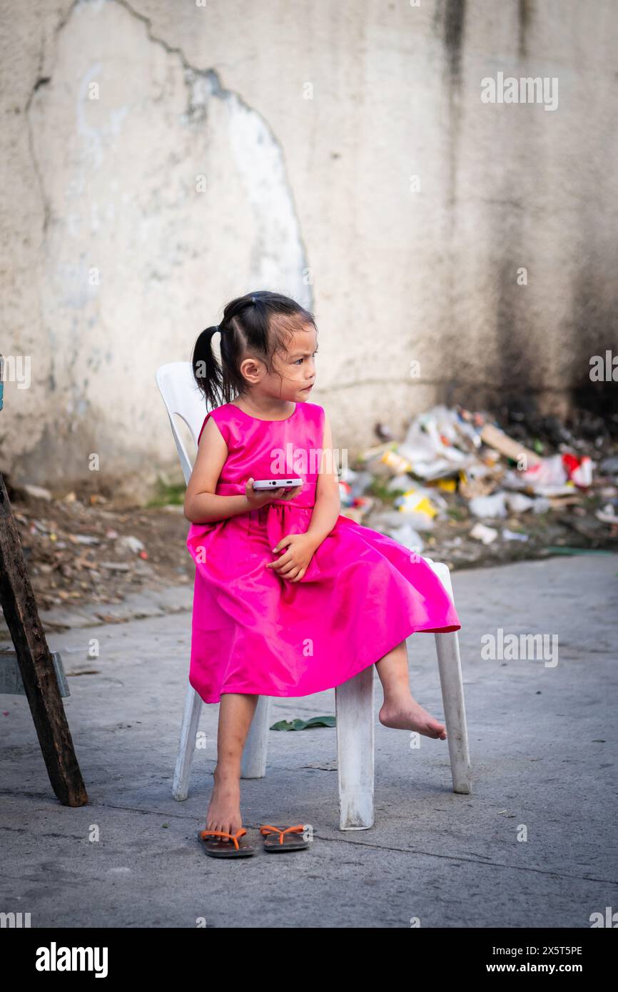 Une jeune fille philippine dans une jolie robe rose regarde son téléphone devant sa maison à Angeles City, aux Philippines. Banque D'Images