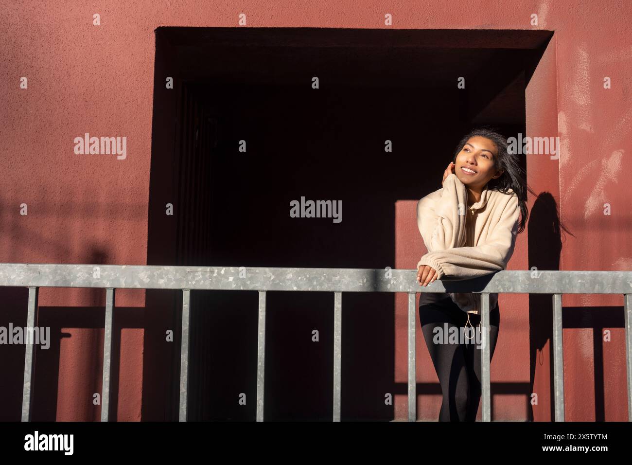 Portrait d'une femme souriante appuyée sur une balustrade contre un mur rouge Banque D'Images