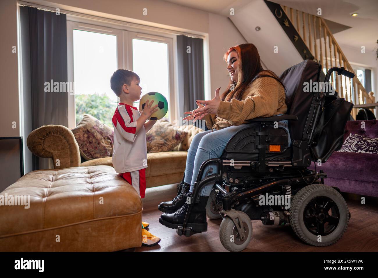 Femme en fauteuil roulant jouant avec son fils en uniforme de football dans le salon Banque D'Images Femme en fauteuil roulant jouant avec son fils en uniforme de football dans le salon Banque D'Images