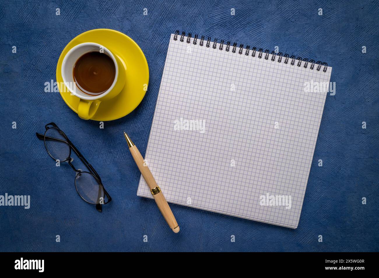 cahier à spirale vierge avec papier ligné, pose à plat avec café, lunettes de lecture et stylo sur papier d'art texturé bleu Banque D'Images