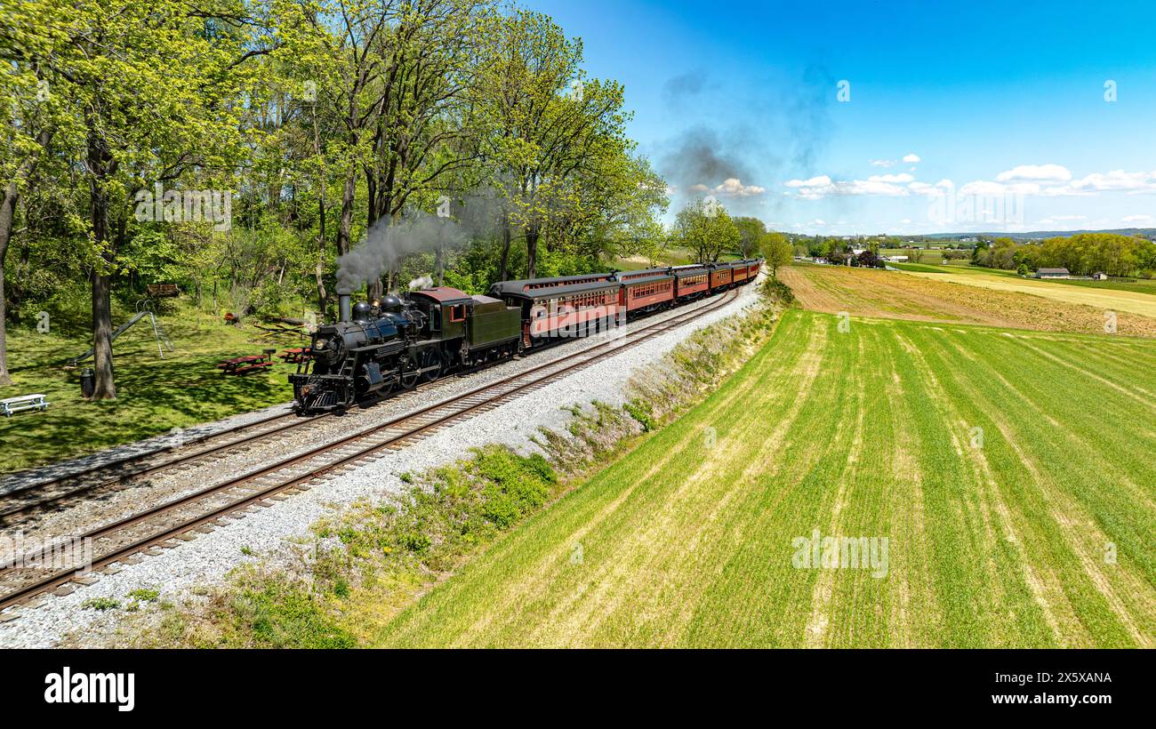 Une vue aérienne enchanteresse d'un train à vapeur historique émettant un nuage de fumée alors qu'il voyage à travers une campagne luxuriante, flanquée d'un champ vert vibrant Banque D'Images