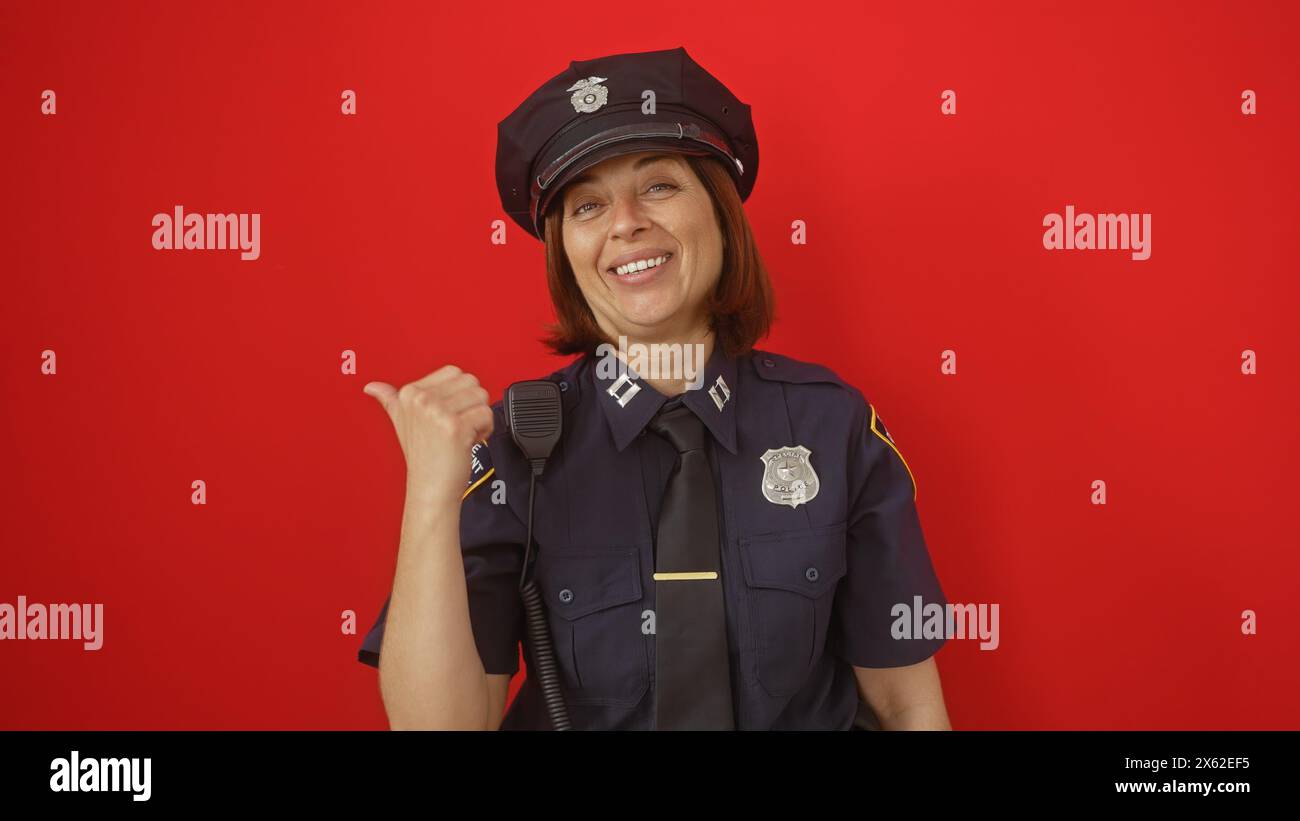 Policier hispanique souriant d'âge moyen faisant des gestes avec le pouce sur un fond rouge. Banque D'Images