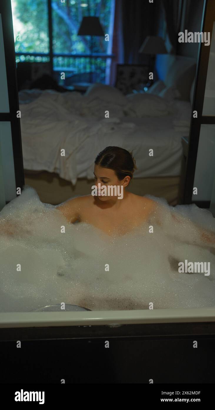 Une jeune femme hispanique se détend dans un bain moussant avec la chambre d'un hôtel de luxe en arrière-plan. Banque D'Images