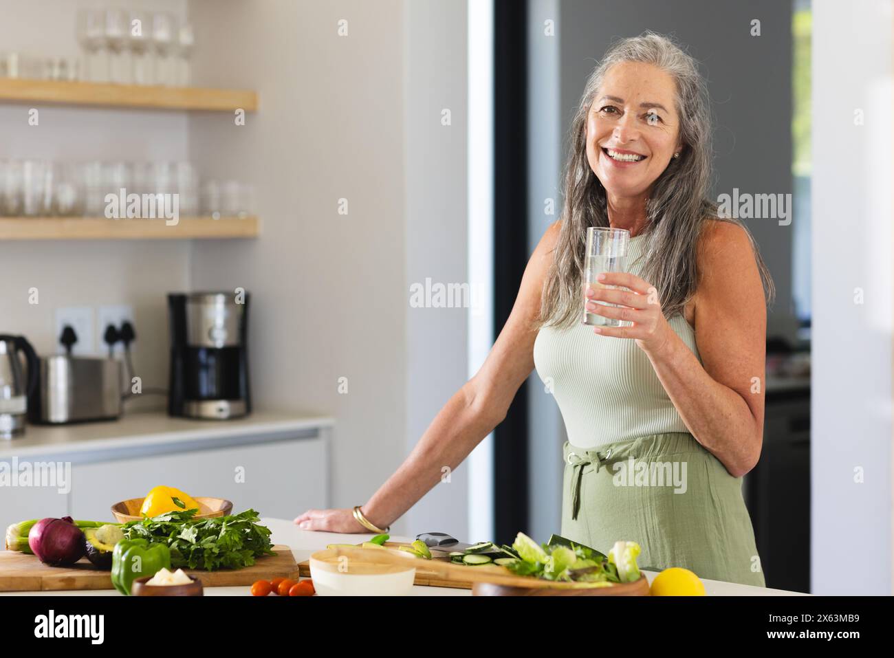 À la maison, femme mature caucasienne tenant un verre d'eau, debout dans la cuisine, espace de copie Banque D'Images