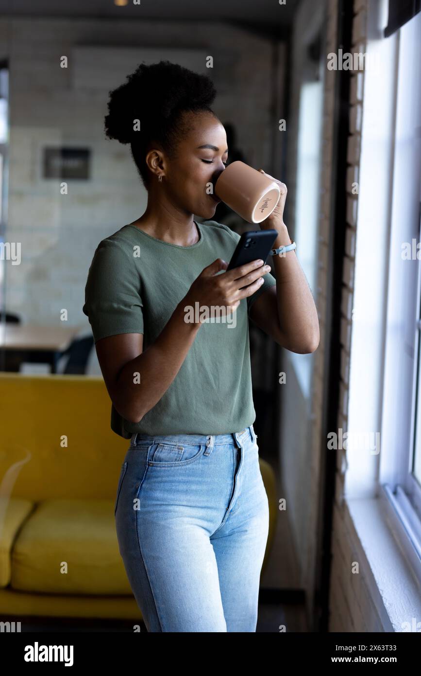 Au bureau, femme d'affaires afro-américaine buvant du café, vérifiant le téléphone Banque D'Images