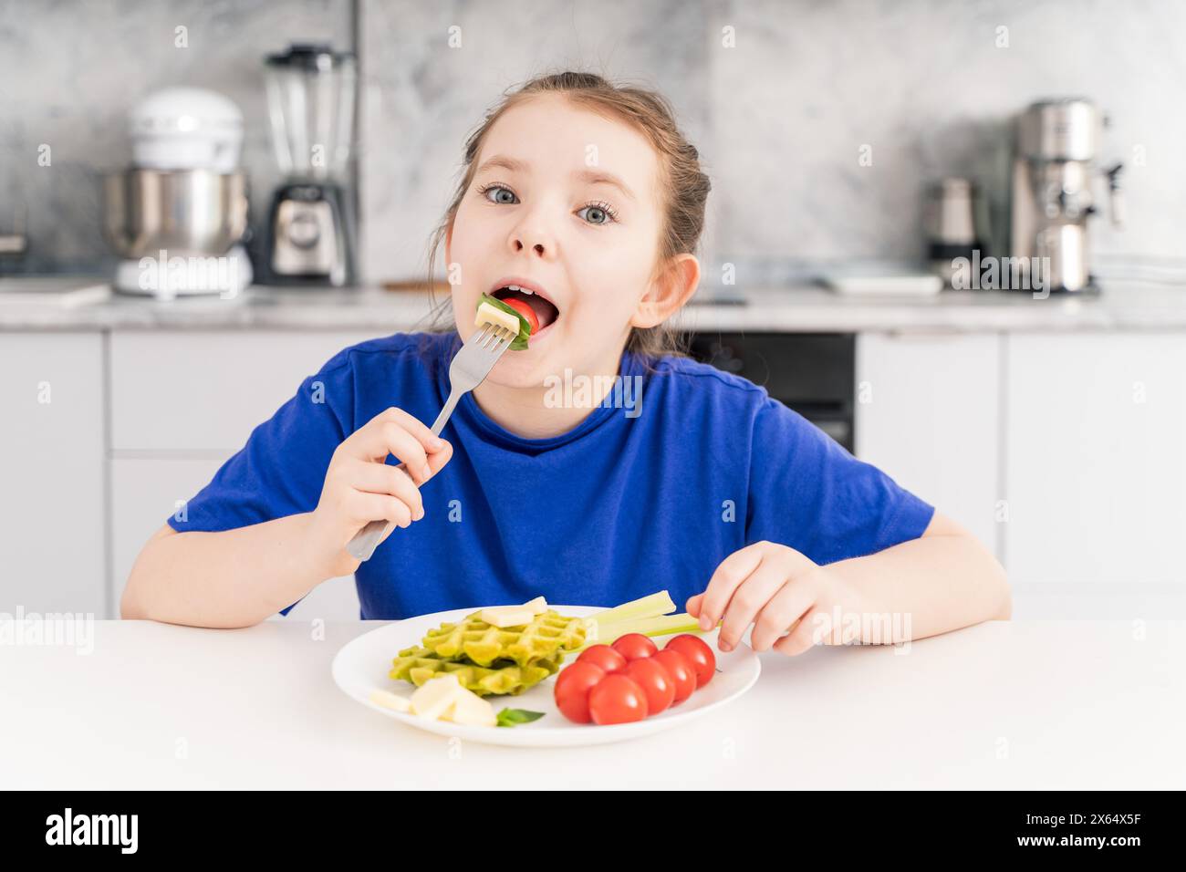 Heureuse petite fille tenant une fourchette dans la bouche. Une jeune fille pré-adolescente prend le petit déjeuner dans la cuisine à la maison avec une assiette de gaufres, légumes et fromage. Banque D'Images