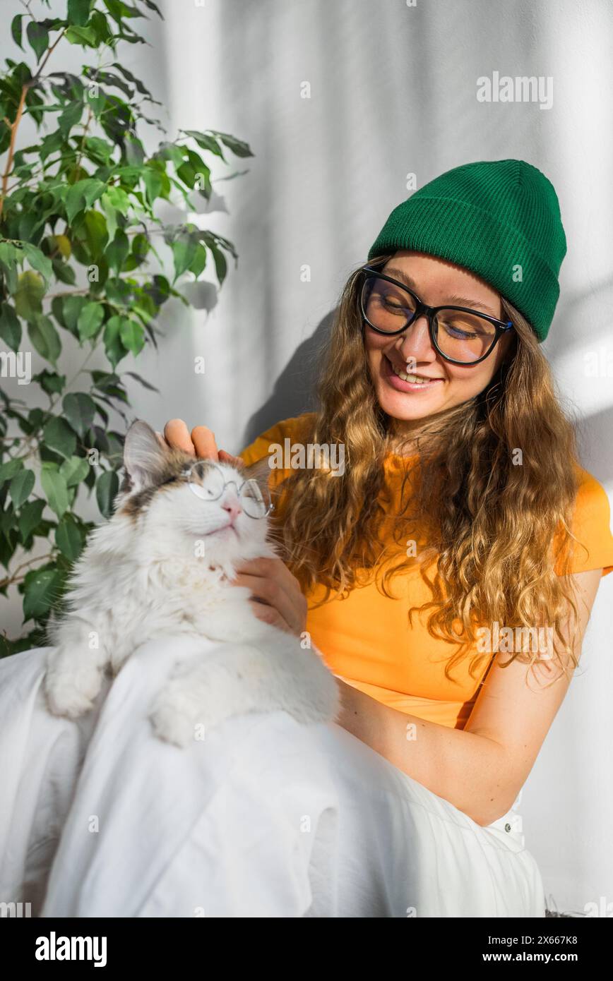 Jeune femme joyeuse dans des lunettes et un bonnet vert caresse avec amour son chat blanc moelleux, tous deux profitant d'un moment confortable ensoleillé ensemble. Banque D'Images