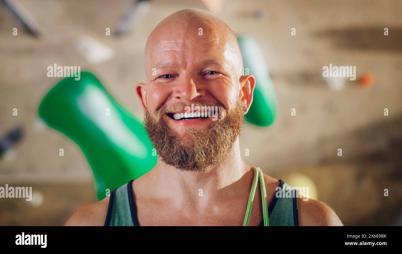 Athlète masculin masculin fort souriant et posant au Rock Climbing Gym avec fond de mur de Bouldering. Beau homme chauve heureux avec Portrait de barbe de gingembre, portant un maillot de corps coloré. Banque D'Images