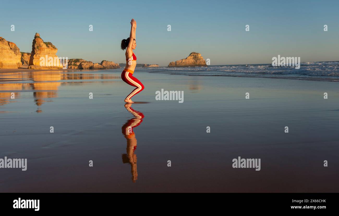 femme faisant chaise de yoga pose sur la plage, reflets dans l'eau. Banque D'Images