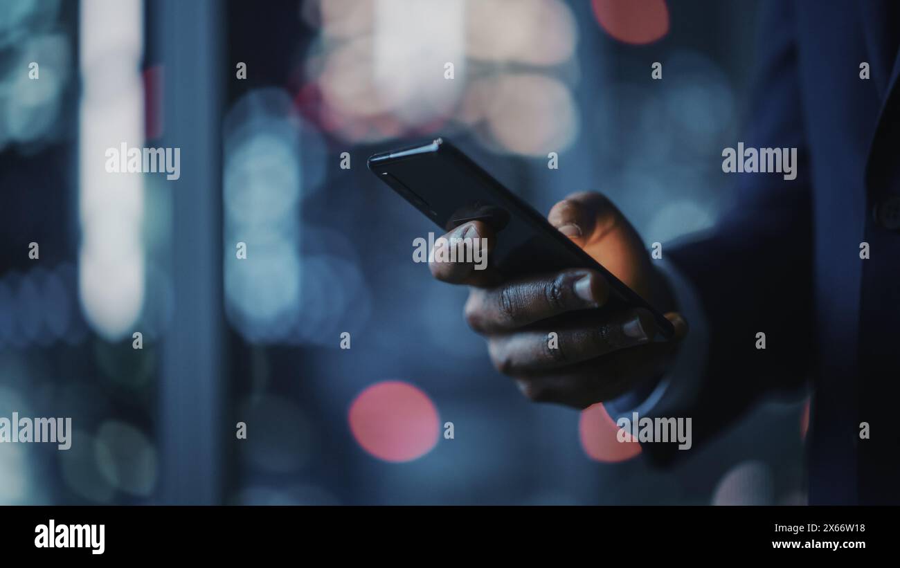 Bureau de nuit : homme d'affaires noir réussi portant costume debout, en utilisant smartphone. CEO navigation sur Internet, gestion de la stratégie des médias sociaux dans le logiciel de commerce électronique. Concentrez-vous sur la main avec le téléphone portable Banque D'Images