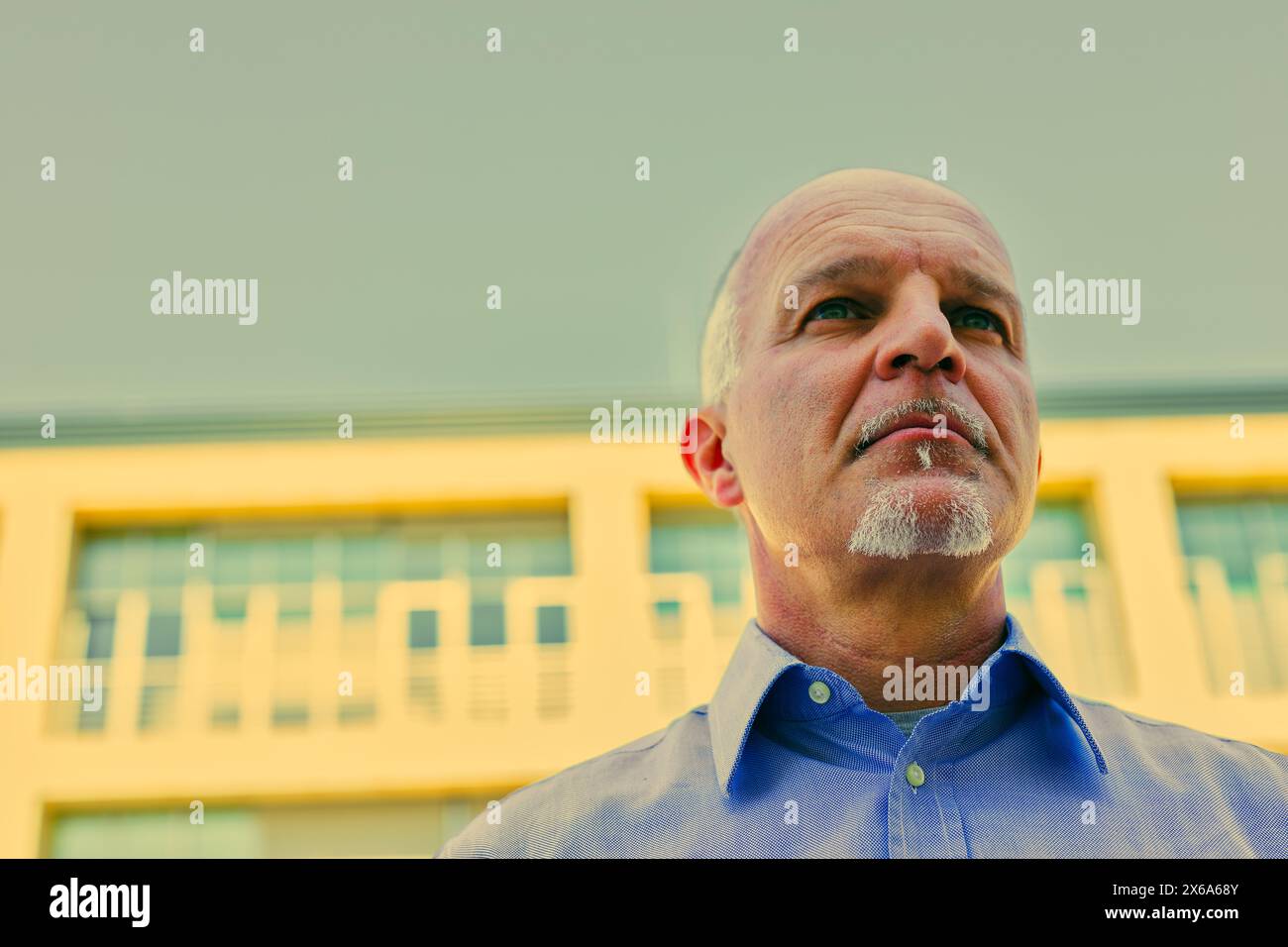 Un homme d'âge mûr réfléchi regarde vers le haut, son expression reflétant la contemplation profonde contre un bâtiment jaune moderne Banque D'Images