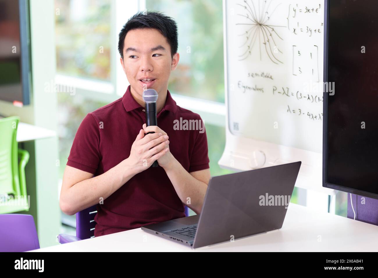 Un étudiant est assis à une table dans la salle de classe, répondant à une question de discussion avec le microphone. Il a un ordinateur portable devant lui. Banque D'Images