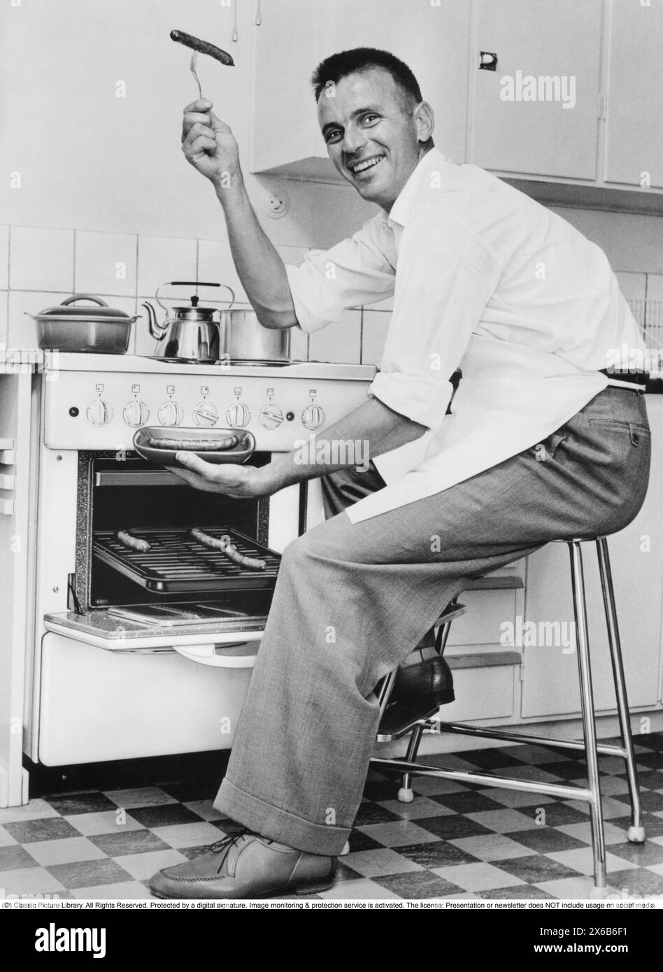Homme dans la cuisine dans les années 1960 Il tient une saucisse prête à l'emploi sur la fourchette. Il a grillé les saucisses au four. Le tabouret de cuisine est un véritable classique du mobilier suédois. Un tabouret AWAB qui a commencé à être fabriqué en 1956 par Anderstorp Werkstad AB et a été fabriqué à plus de 70 000 exemplaires. On l'appelait parfois la chaise de la femme au foyer. Banque D'Images