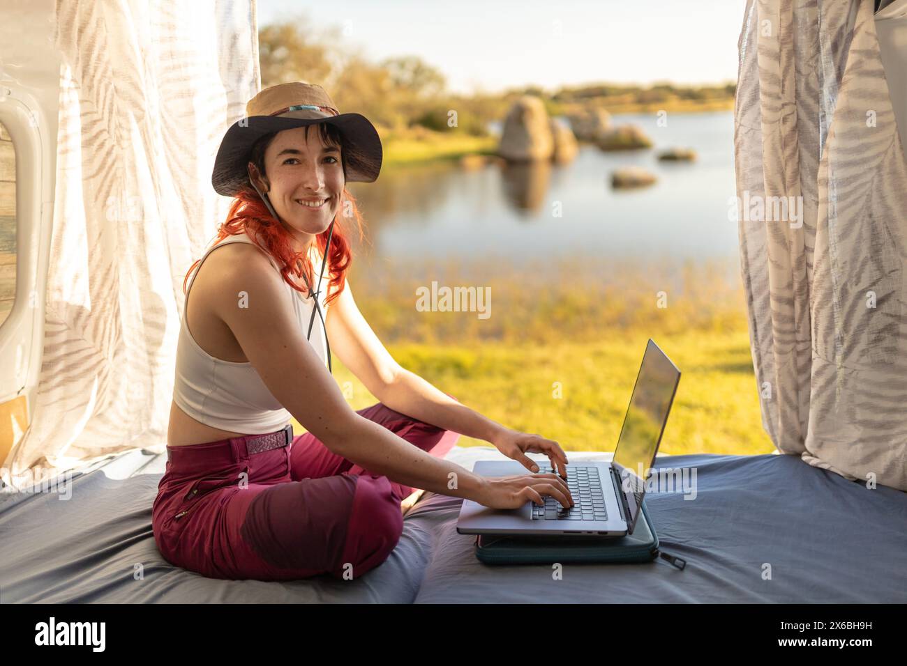 Femme aux cheveux rouges télétravail tapant avec ordinateur portable dans le camping-car au milieu de la nature près du lac, connecté à Internet, regardant la caméra et souriant, Banque D'Images