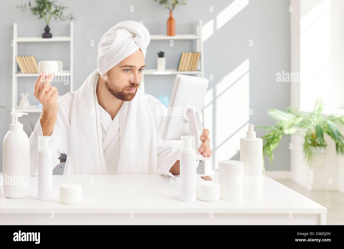 Homme drôle en peignoir blanc et serviette assis à table avec des bouteilles cosmétiques ayant la journée de beauté Banque D'Images