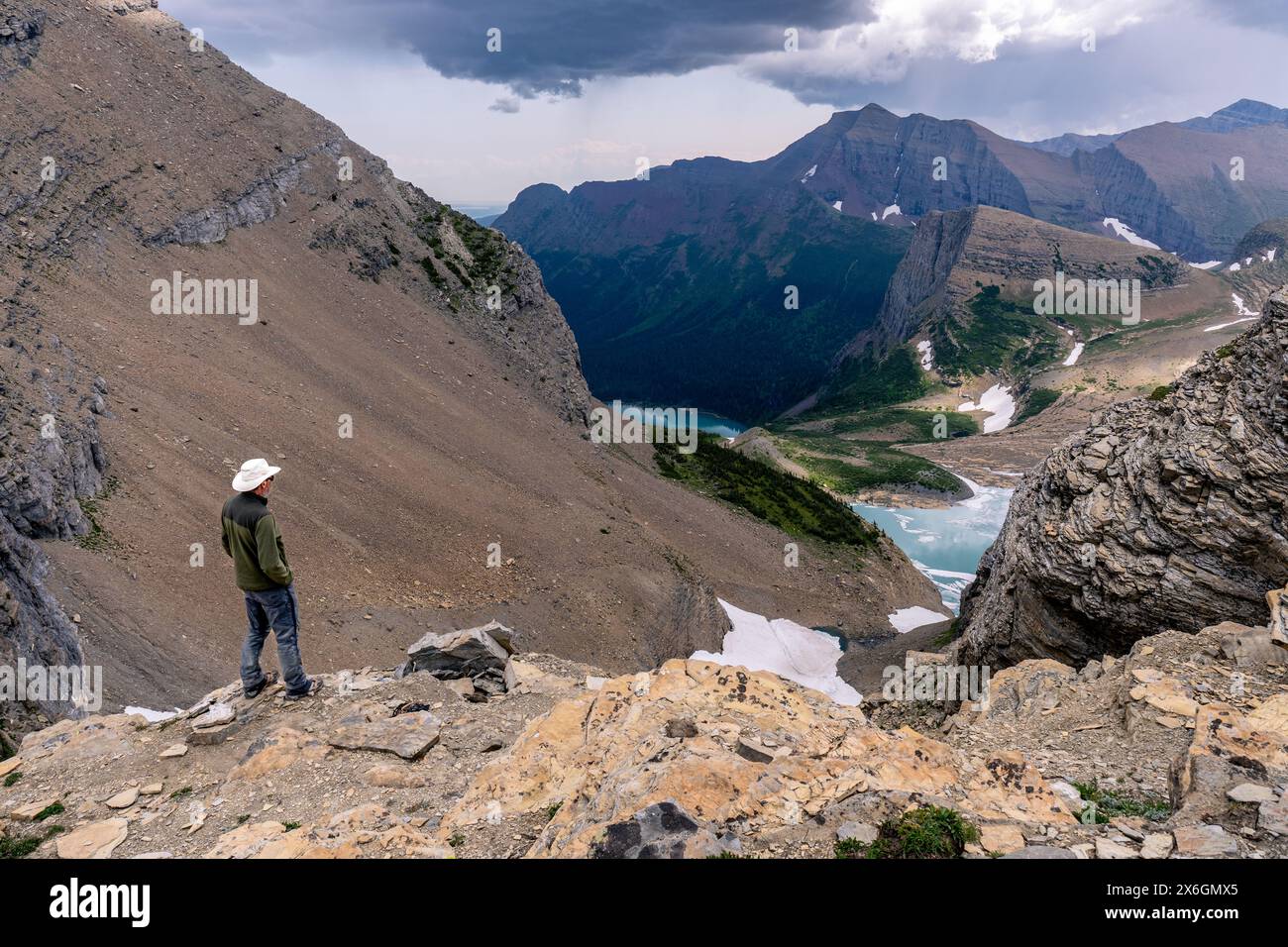 Homme caucasien mûri debout au bord d'une falaise regardant une scène d'un lac glaciaire dans un canyon profond avec des taches de neige et de hautes montagnes Banque D'Images