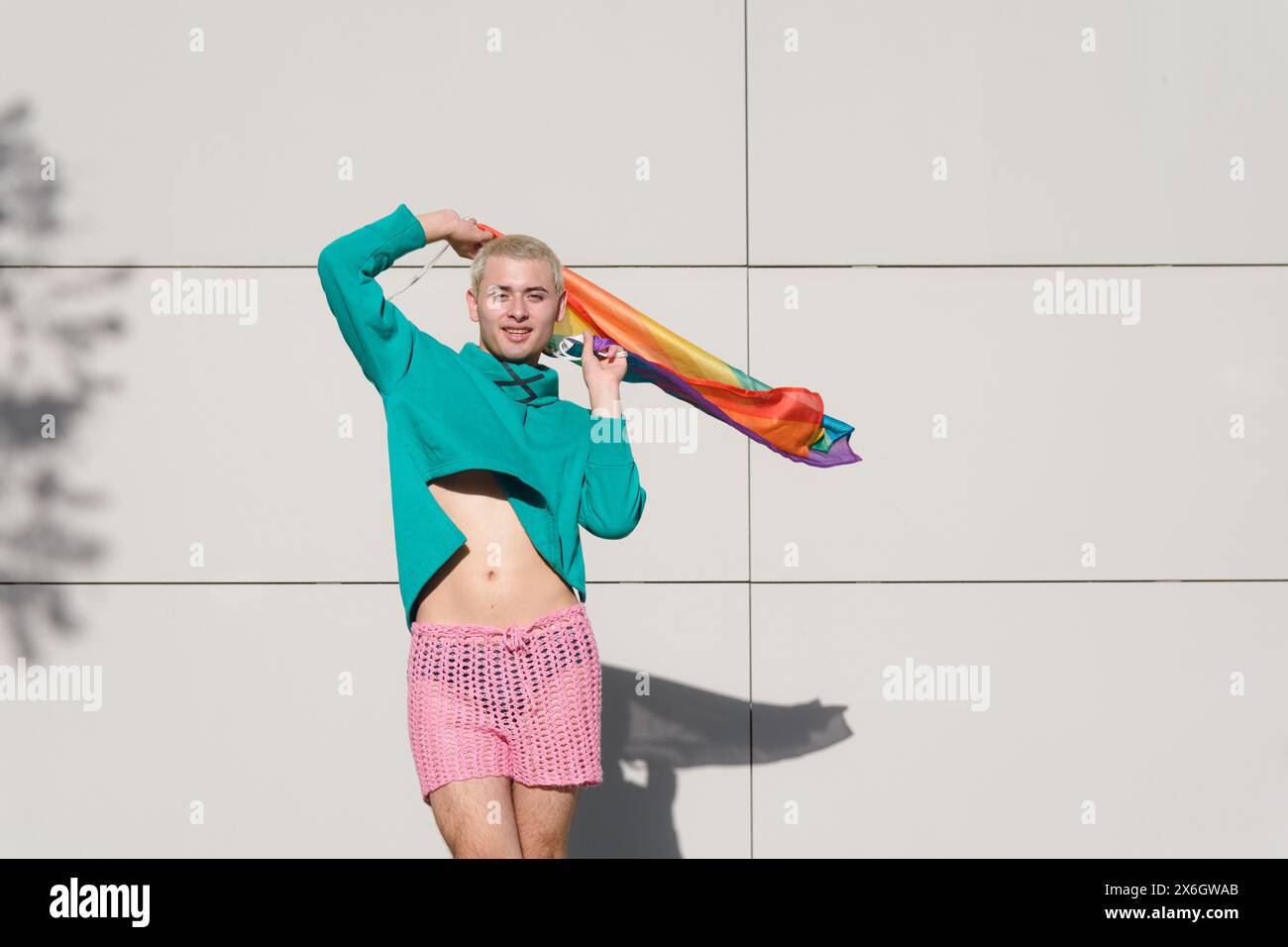 jeune homme latino blond aux cheveux courts portant un pull vert et un short rose dansant avec drapeau de fierté à l'extérieur, il est très fier d'être gay. Banque D'Images