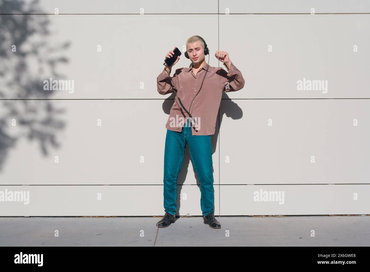 jeune homme latin blond aux cheveux courts à l'extérieur au coucher du soleil avec chemise brune et pantalon jean avec écouteurs dansant et écoutant de la musique, il est agréable Banque D'Images