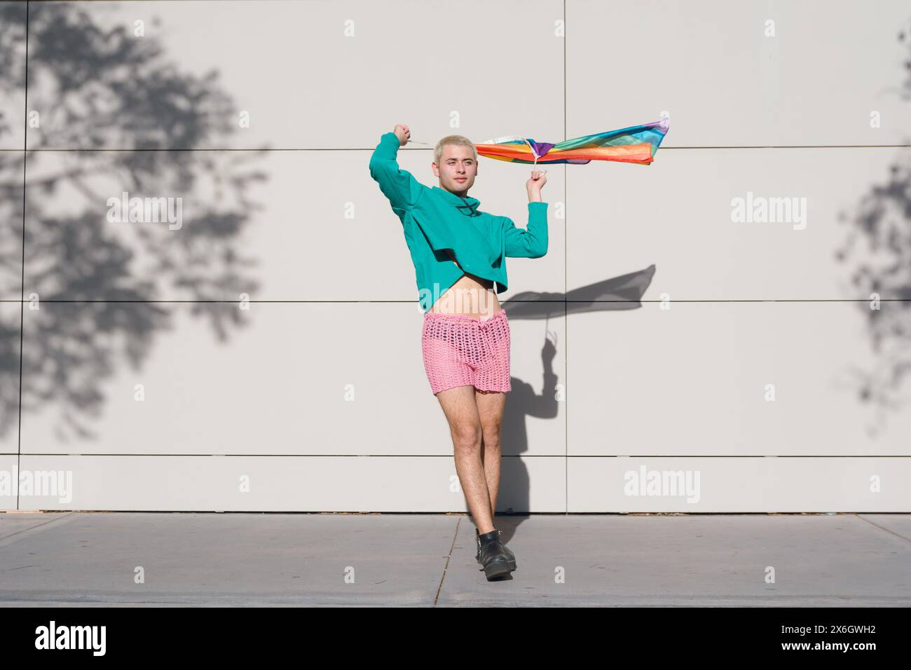 Jeune homme latin aux cheveux blonds courts, portant un short rose, des baskets noires et un pull vert, est à l'extérieur avec le drapeau de fierté dansant et profitant du coucher du soleil Banque D'Images