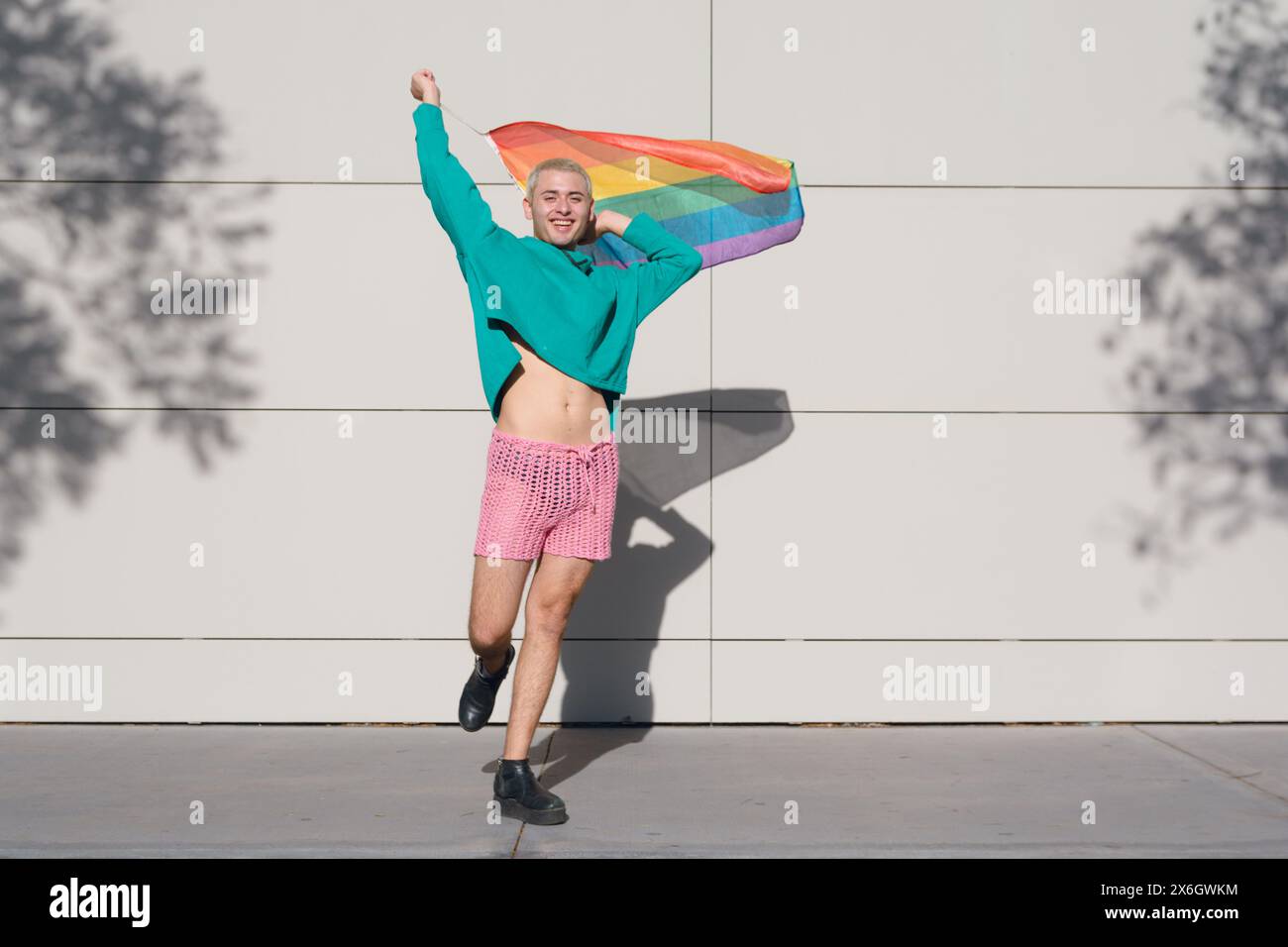 Jeune homme latin aux cheveux blonds courts, portant un short rose, des baskets noires et un pull vert, est à l'extérieur avec le drapeau de fierté dansant et profitant du coucher du soleil Banque D'Images