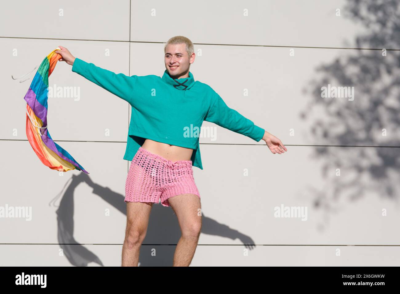 jeune homme latino blond aux cheveux courts portant un pull vert et un short rose dansant avec drapeau de fierté à l'extérieur, il est très fier d'être gay. Banque D'Images