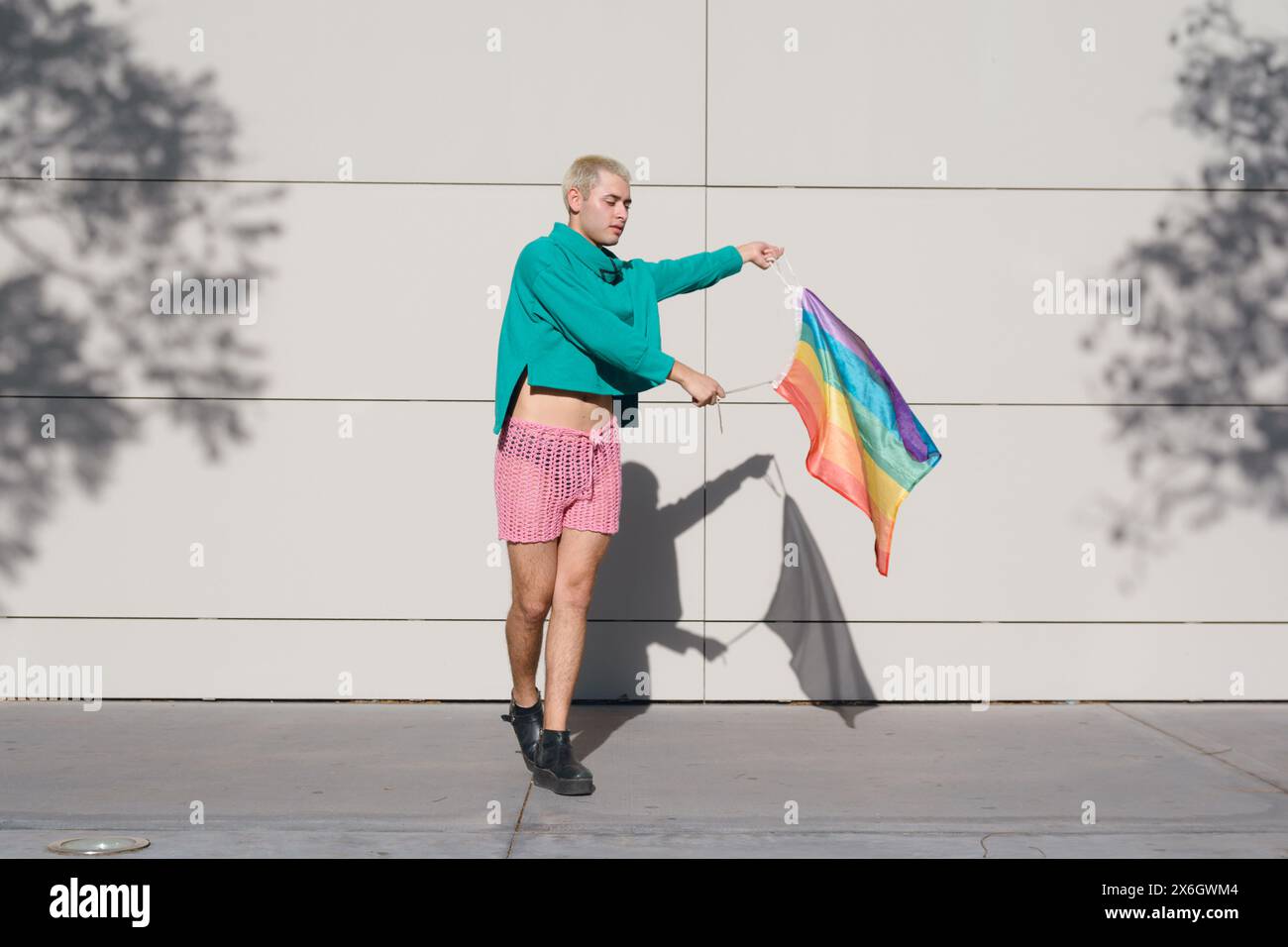 Jeune homme latin aux cheveux blonds courts, portant un short rose, des baskets noires et un pull vert, est à l'extérieur avec le drapeau de fierté dansant et profitant du coucher du soleil Banque D'Images