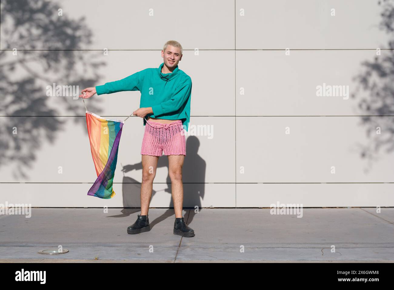 Jeune homme latin aux cheveux blonds courts, portant un short rose, des baskets noires et un pull vert, est à l'extérieur avec le drapeau de fierté dansant et profitant du coucher du soleil Banque D'Images