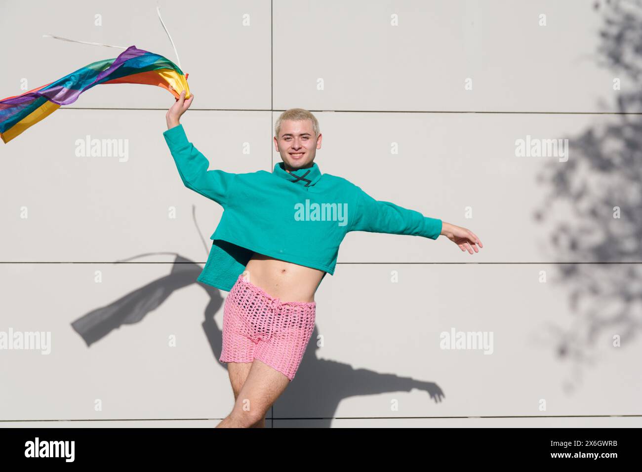jeune homme latino blond aux cheveux courts portant un pull vert et un short rose dansant avec drapeau de fierté à l'extérieur, il est très fier d'être gay. Banque D'Images