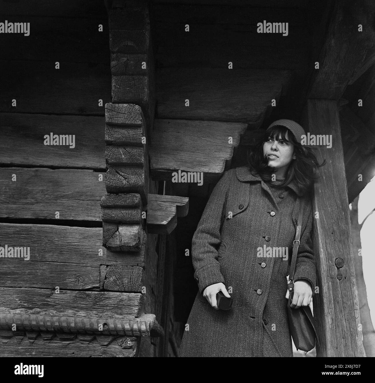 Roumanie, approx. 1973. Jeune femme en tenue d'hiver chic posant par une vieille maison en bois. Banque D'Images