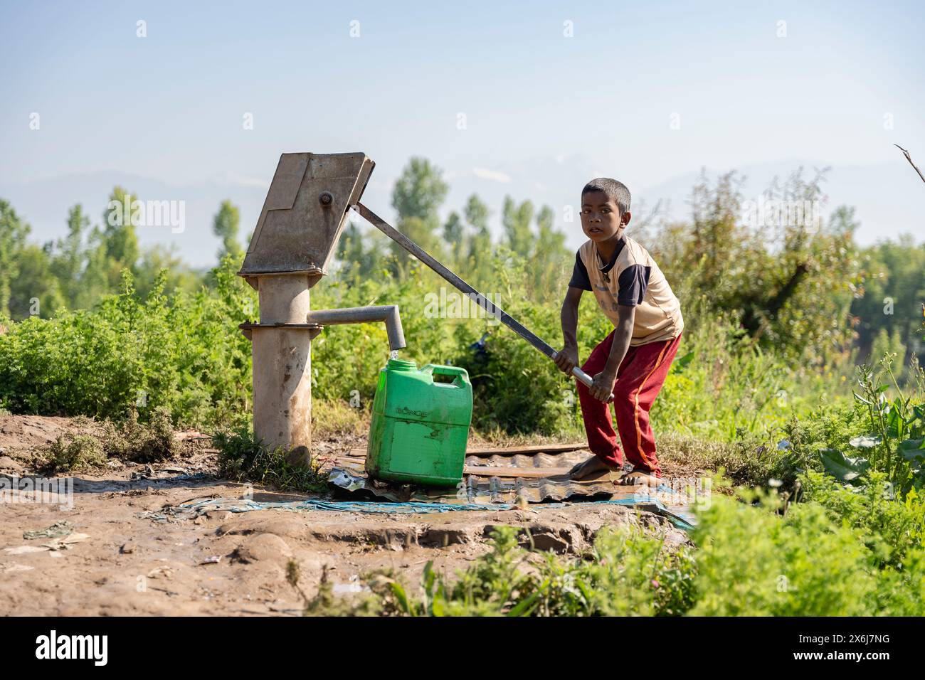 Un garçon non local remplit un récipient avec de l'eau d'une pompe à main par une journée ensoleillée. Banque D'Images