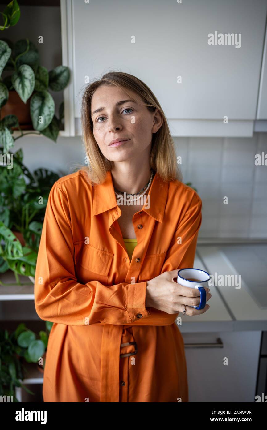 Femme heureuse détendue debout à la cuisine de la maison tenant la tasse regardant l'entourage de la caméra par des plantes d'intérieur Banque D'Images