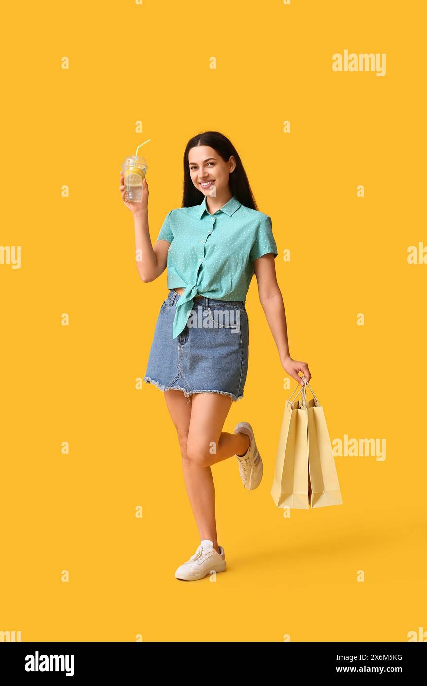 Jeune femme heureuse avec tasse de limonade fraîche et sacs à provisions sur fond jaune Banque D'Images