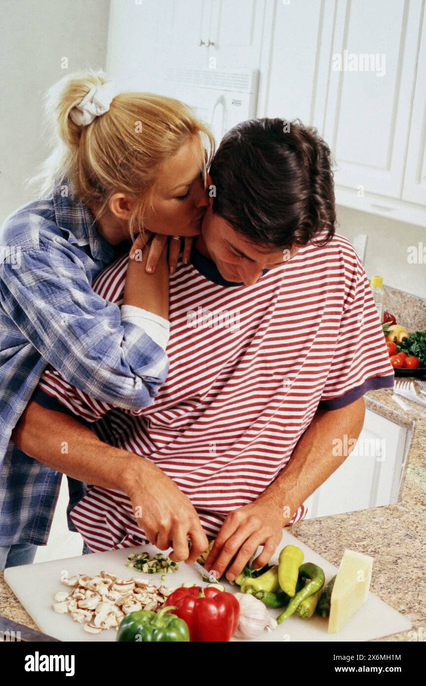 Un homme et une femme s'embrassent passionnément tout en étant engagés dans la tâche de hacher des légumes dans un cadre de cuisine confortable. Banque D'Images