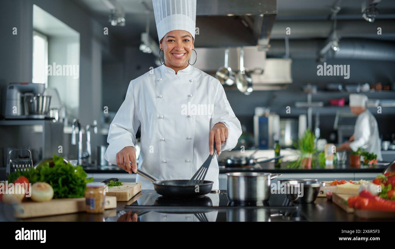 Cuisine du restaurant : Portrait de la femme chef noire en action, utilise Pan pour cuisiner délicieux, traditionnel authentique nourriture, regarde l'appareil photo et sourires. Plats gastronomiques sains. Grand angle moyen Banque D'Images