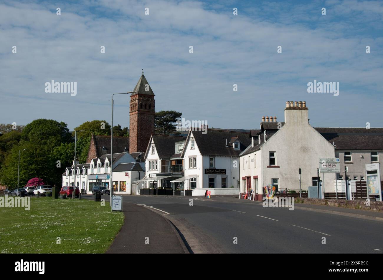 Main Street, Lamlash, Île d'Arran, Écosse, Royaume-Uni - magasins, maisons, église, clocher d'église ; tour d'église ; vert village. jolies maisons blanches Banque D'Images
